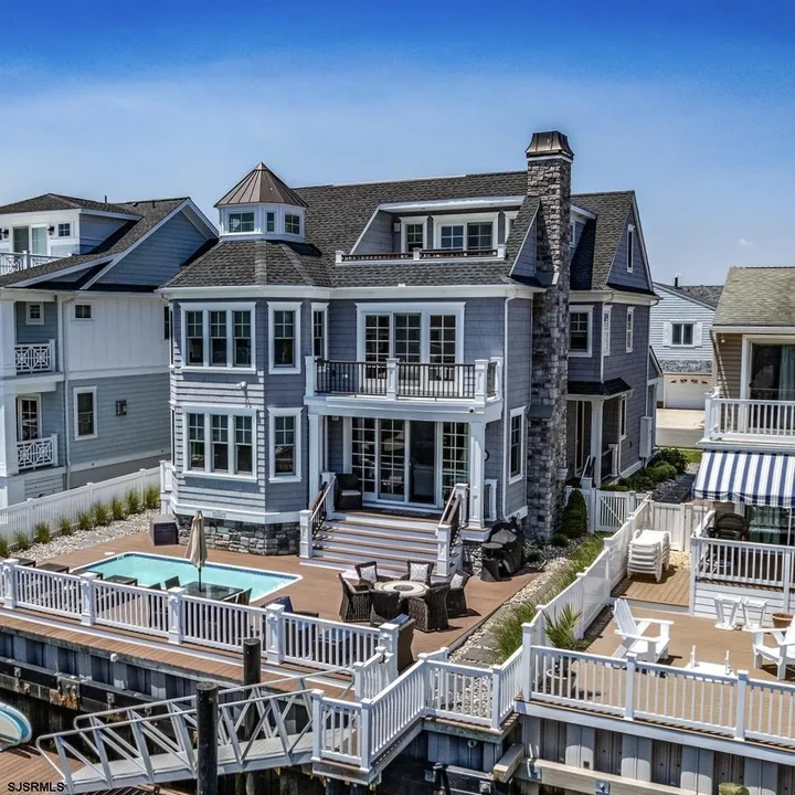 We see the front of a blue house in Ocean City, NJ against a clear blue sky. The house has a pool in the front flanked by several sets of tables and chairs.