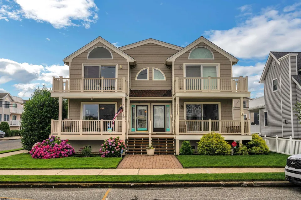 We see the front of a beige and brown house in Ocean City, NJ adorned by green flora and clean-cut grass against a clear blue sky.